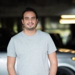 A man in a gray t - shirt standing in front of a car.